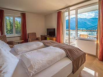 Bright bedroom with a large bed, chairs, and balcony doors overlooking mountains and trees in sunlight.