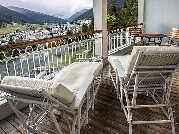 White wicker lounge chairs and a small table on a wooden balcony overlooking a mountain town and valley