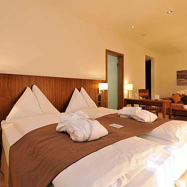 A modern hotel bedroom with a large bed, wooden headboard, bedside lamps, and seating area in warm light.