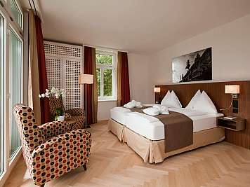 Modern hotel bedroom with twin beds, patterned armchairs, large windows, and warm wood accents.