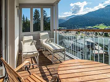 Sunny balcony with two lounge chairs, a wooden table, and mountain views beyond glass railings.