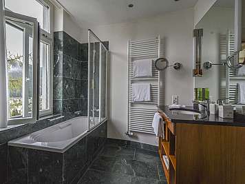 Bright bathroom with a bathtub, glass shower, vanity, and open window overlooking mountains and trees.