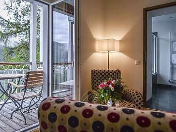 Warm interior with patterned chairs, a floor lamp, flowers, and an open balcony door to a mountain view.