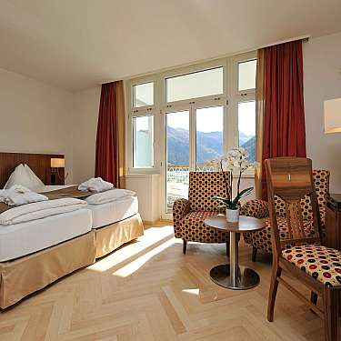 Bright hotel room with twin beds, patterned chairs, and mountain view windows