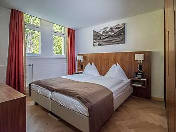 Aufgeräumtes Hotelzimmer mit Doppelbett, Holzpaneel, roten Vorhängen und Blick ins Grüne am Fenster, 