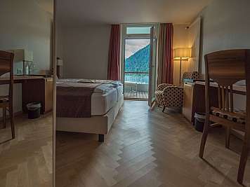 A tidy hotel room with twin beds, desks, patterned chairs, and an open balcony door overlooking mountains.