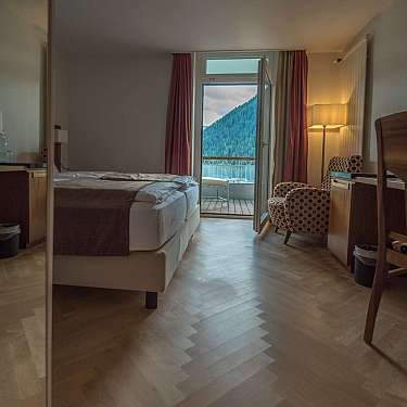 Neat hotel room with twin beds, wooden desks, and an open balcony door to a mountain view.