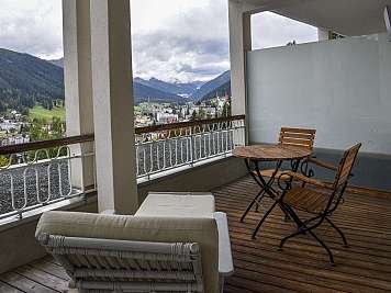 Wooden balcony with table and chairs overlooking a mountain town and cloudy valley landscape.