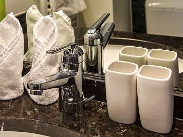 Chrome faucet on a dark stone bathroom counter with folded white towels and ceramic cups nearby, reflected in a mirror, 