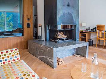 Modern living room with a central fireplace, wood flooring, desk, chair, and patterned sofa.