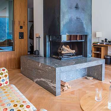 Moderner Wohnraum mit zentralem Kamin, Holzverkleidung, Schreibtisch und gepunkteter Polsterbank in warmen Holztönen. Zum Inhalt