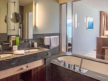 Modern bathroom with double sinks, mirrors, wall lights, and a bathtub beside a glass partition for the bedroom area.
