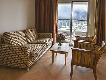 Living room with patterned sofa, two wooden armchairs, coffee table, and balcony mountain view.