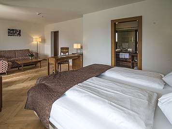 Spacious hotel suite with a bed, desk, sofa, lamps, and an open bathroom doorway.,
