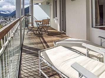 Sunlit apartment balcony with cushioned lounge chairs, small table, and town and mountains beyond the railing.