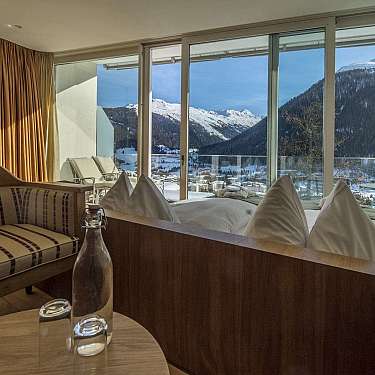 Cozy hotel room with a bed, sofa, and balcony overlooking snowy mountains and a valley.