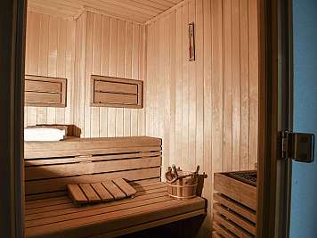 Innenraum einer Holzsauna mit Sitzbänken, Kopfstütze, Eimer und Thermometer an der Wand, gesehen durch eine geöffnete Tür; true}
