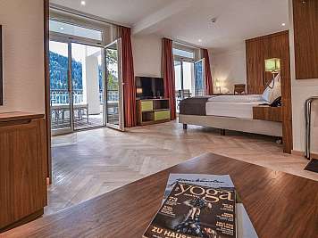 Modernes Hotelzimmer mit Bett, Fernseher und offenem Balkonblick auf Berge, auf dem Tisch liegt eine Zeitschrift, content_suitable: true