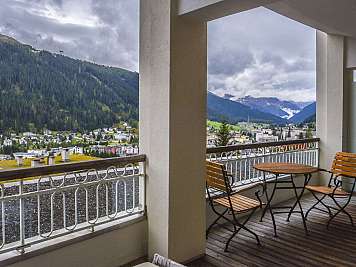 Überdachter Balkon mit Holztisch und zwei Stühlen, Blick auf Bergtal und Ort unter wolkenverhangenem Himmel, true},