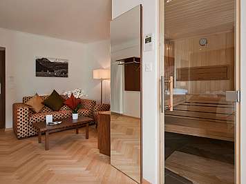 Gemütlicher Wohnbereich mit gemustertem Sofa, Couchtisch und Blick in eine Holzsauna durch Glastür, 