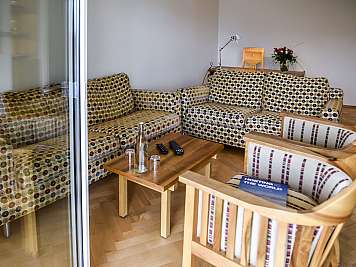 Bright lounge with patterned sofas, wooden chairs, coffee table, and desk by the wall with flowers and lamp