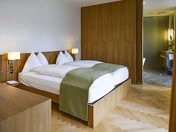 Modern hotel bedroom with a wooden bed, white bedding, bedside lamps, and an open bathroom area in the background.