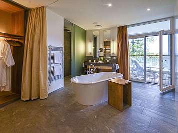 Spacious modern bathroom with freestanding tub, vanity, robe alcove, and open balcony doors to lounge chairs on a deck.