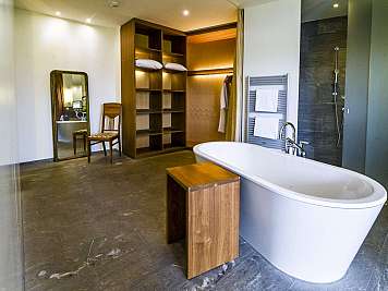 Spacious modern bathroom with a white freestanding bathtub, wood shelving, mirror, and shower area with towels and dark tile walls, true}