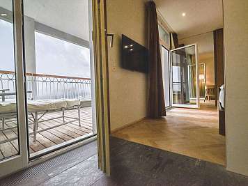Bright hotel room with open balcony doors, wall-mounted TV, and a view of the terrace seating outside.
