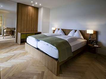 A modern hotel room with a double bed, bedside lamps, wooden flooring, and a sitting area in the background.