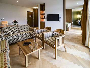 Modern hotel suite with patterned sofa, armchairs, coffee table, wall-mounted TV, and bedroom visible beyond curtains