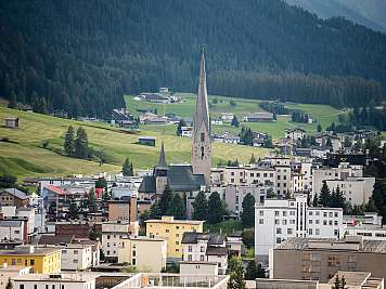 Alpines Dorf mit hoher Kirche, Wohnhäusern und grünen Hängen vor einem Waldhang. Abgeschiedenes Dorf in den Bergen
