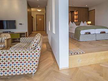 Heller Hotelraum mit Sitzbereich, Fernseher und separatem Schlafbereich mit großem Bett und Holzmöbeln, content_suitable=true}