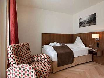 A hotel room with a neatly made bed, patterned armchair, wall lamp, and framed landscape print. The room has wood paneling and red curtains by the window, content_suitable:true