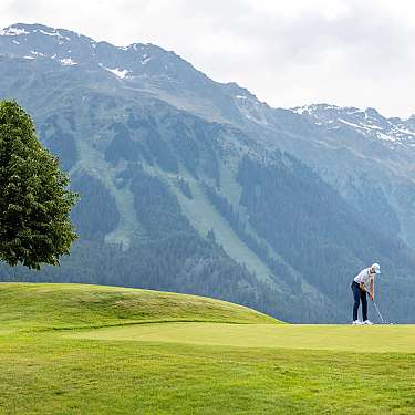 Ein Golfer puttet auf einem gepflegten Grün vor Bergkulisse und Baum.