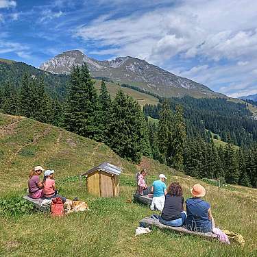 Wandernde sitzen auf einer Almwiese mit Blick auf bewaldete Berge und einen Himmel mit Wolken.