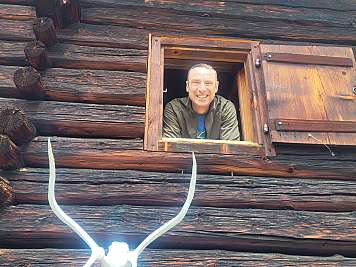 Smiling man leans out of a wooden cabin window above a mounted antler skull.