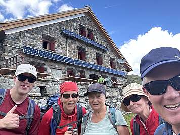 Five hikers posing in front of a stone mountain hut under a blue sky.