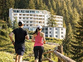 Zwei Personen joggen auf einem Waldweg vor einem großen Hotelgebäude.