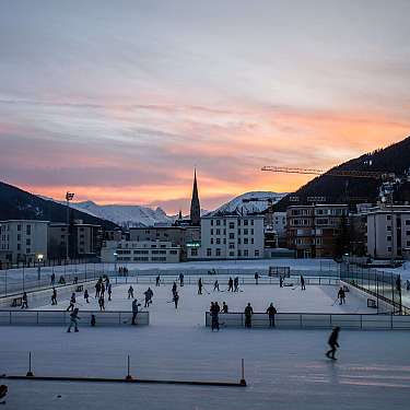 Menschen laufen auf einer Eisbahn vor verschneiten Bergen und einem Sonnenuntergang.