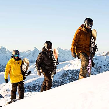 Drei Snowboarder mit Helmen stehen auf einem verschneiten Berggipfel.