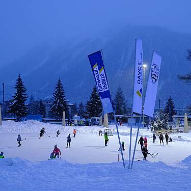 Kinder spielen auf einem beleuchteten Außen-Eislaufplatz im Schnee.