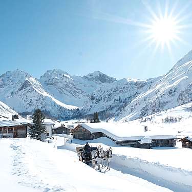 Pferdeschlitten fährt durch verschneites Dorf vor Bergen im Sonnenschein.