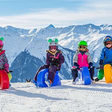 Vier Kinder sitzen mit Helmen auf bunten Skidoo-Schlitten im Schnee vor Bergen.