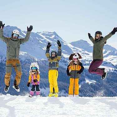 Familie in Skikleidung springt vor verschneiter Bergkulisse.