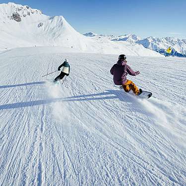 Zwei Wintersportler fahren auf einer breiten, sonnigen Skipiste mit Bergpanorama.