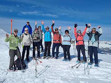 Group of hikers in winter gear stands on a snowy mountain ridge with skis and poles, raising their hands.