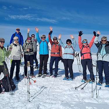 Gruppe von Schneeschuhwanderern steht auf einem verschneiten Berggipfel und winkt in die Kamera.