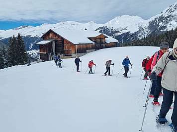Group of hikers snowshoeing past a wooden cabin in a snowy mountain landscape.