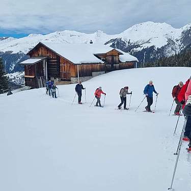 Gruppe von Schneeschuhwanderern auf einer verschneiten Bergwiese vor einer Holzhütte.